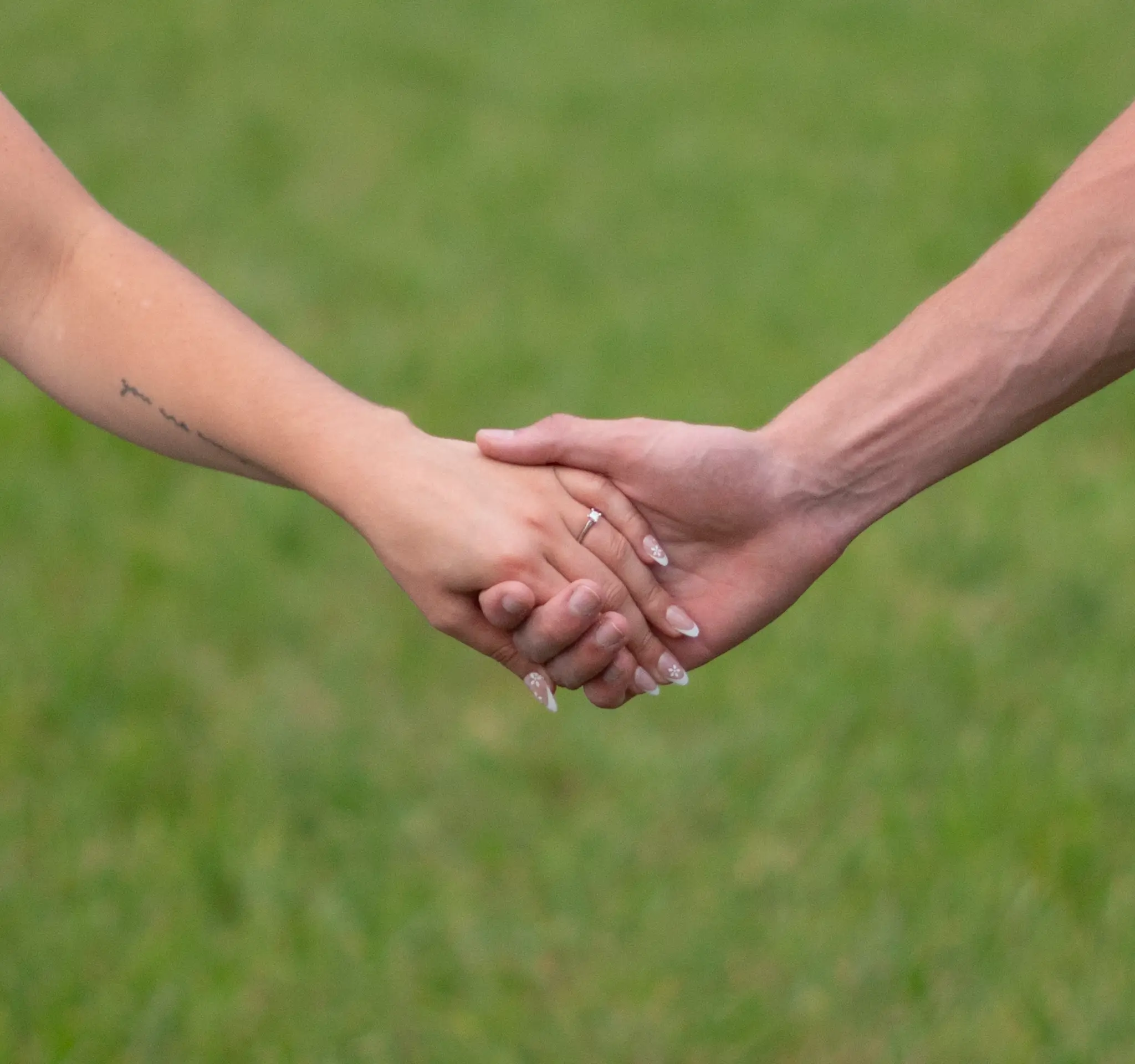 The couple’s hands clasped together over summer grass, engagement ring catching the light
