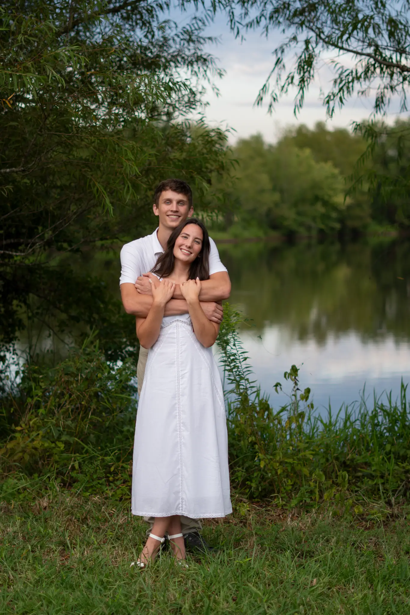 The couple standing together at the water’s edge under tall trees